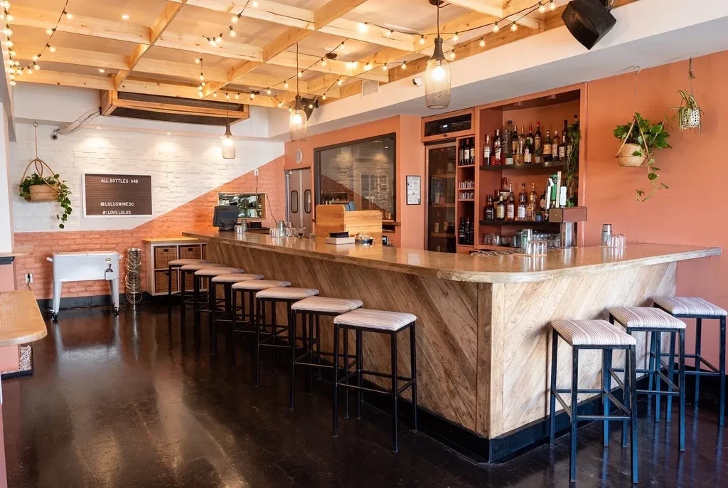 Modern bar interior with chevron wood bar, stocked shelves, and string lights.