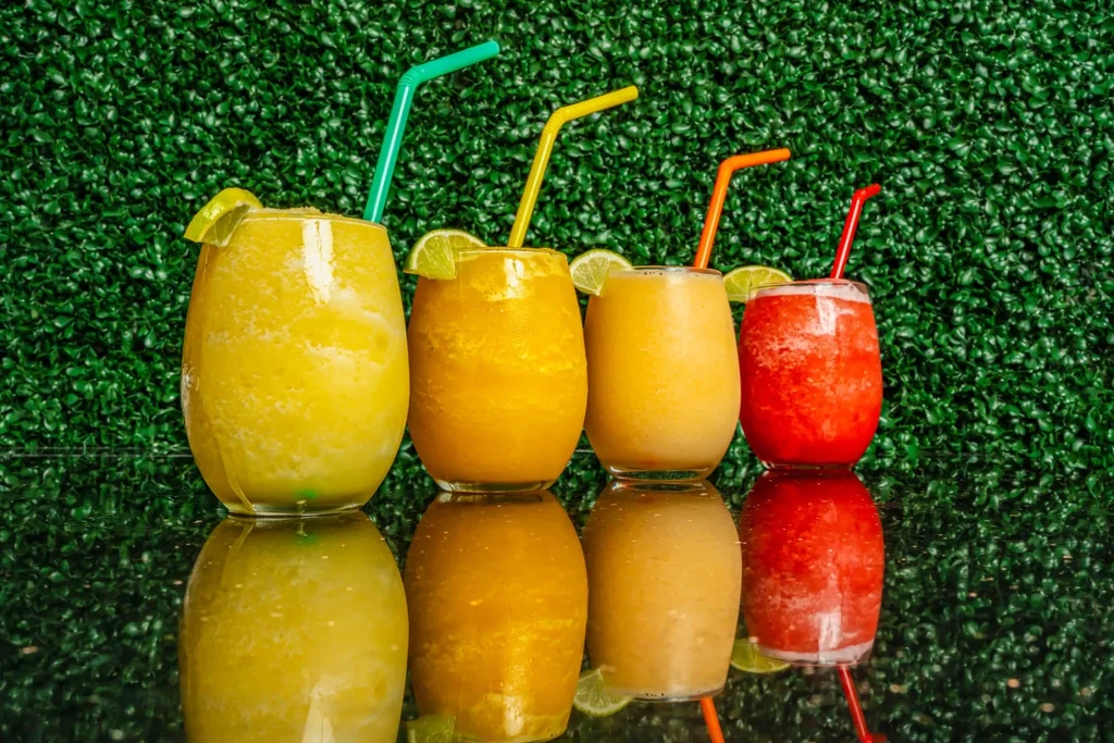 Four colorful frozen drinks with lime and straws on reflective surface.