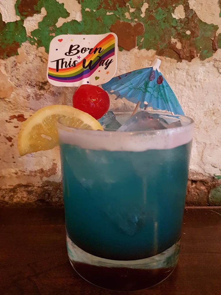 Blue-green cocktail, lemon, cherry, umbrella, and rainbow "Born This Way" pick.