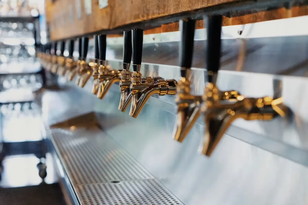 Numerous brass beer taps on a wooden beam above a steel counter.
