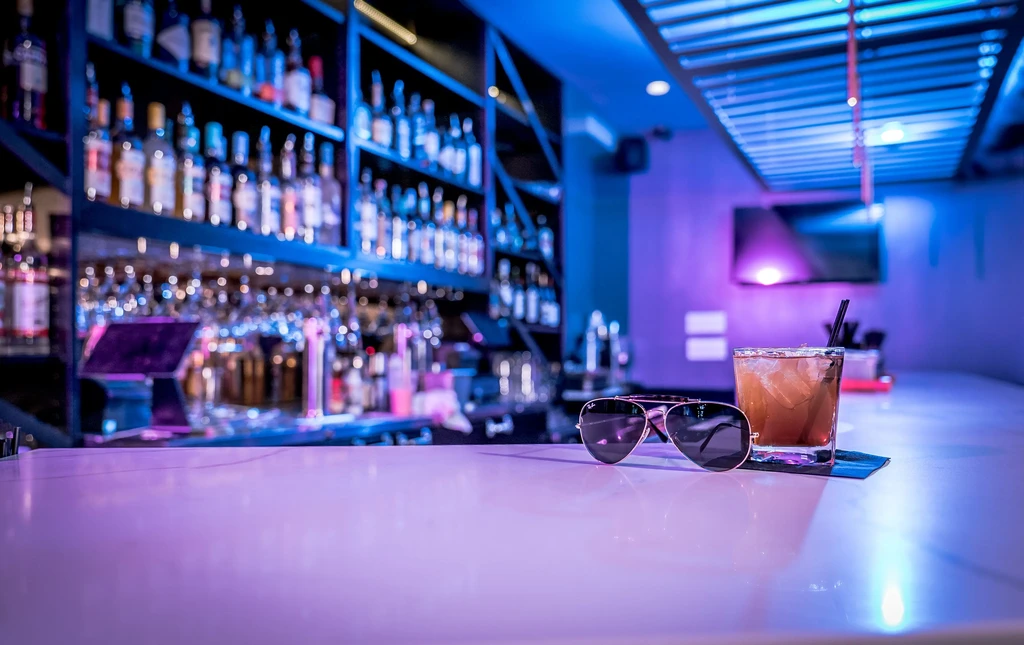 Amber cocktail and sunglasses on a bar counter in purple light.