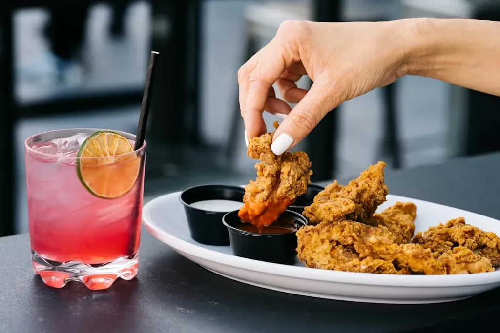 Hand dips golden fried chicken tenders into sauce next to a pink cocktail.