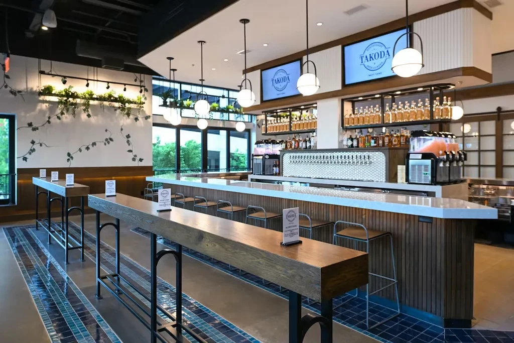 Well-lit modern bar interior with "TAKODA" screens, beer taps, and tables.
