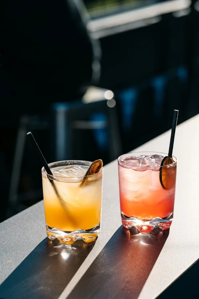 Two vibrant yellow and pink cocktails in wavy tumblers, sunlit.