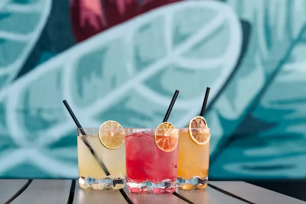 Three colorful cocktails with citrus slices and straws on a wooden table.