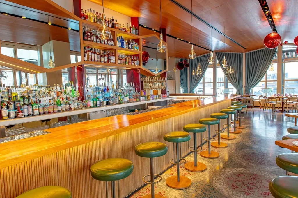 Stylish, empty bar interior with long wooden counter, green stools, liquor display.