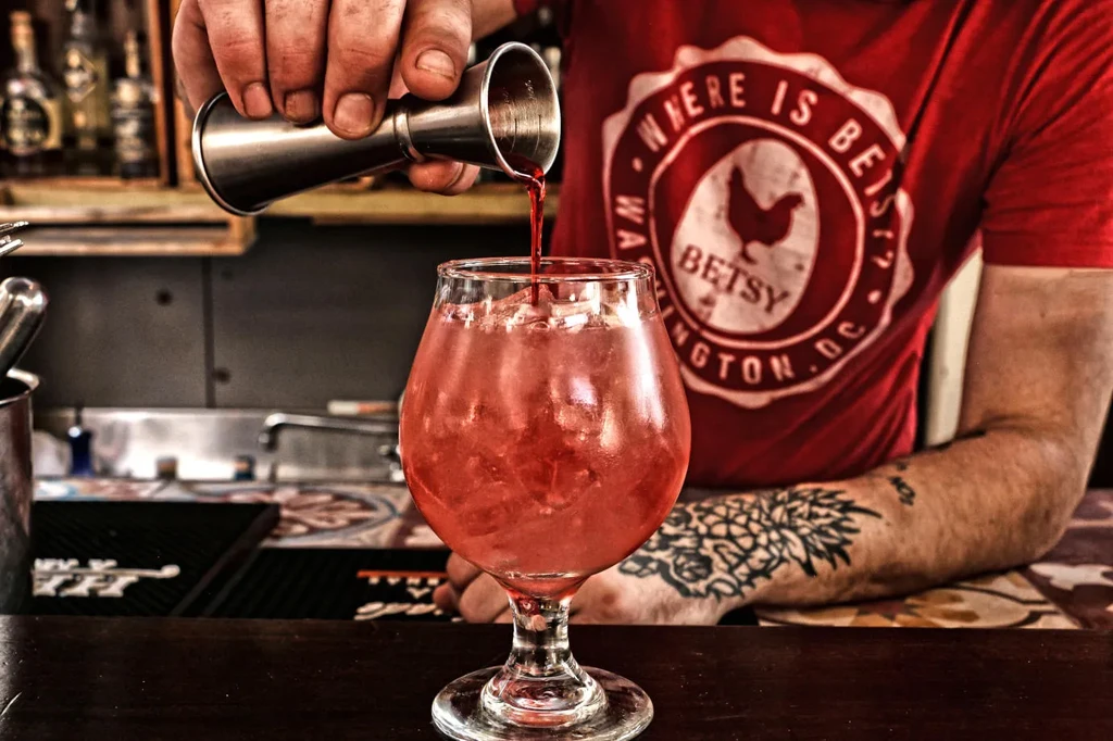 Bartender's tattooed hands pour crimson drink into a glass with ice.