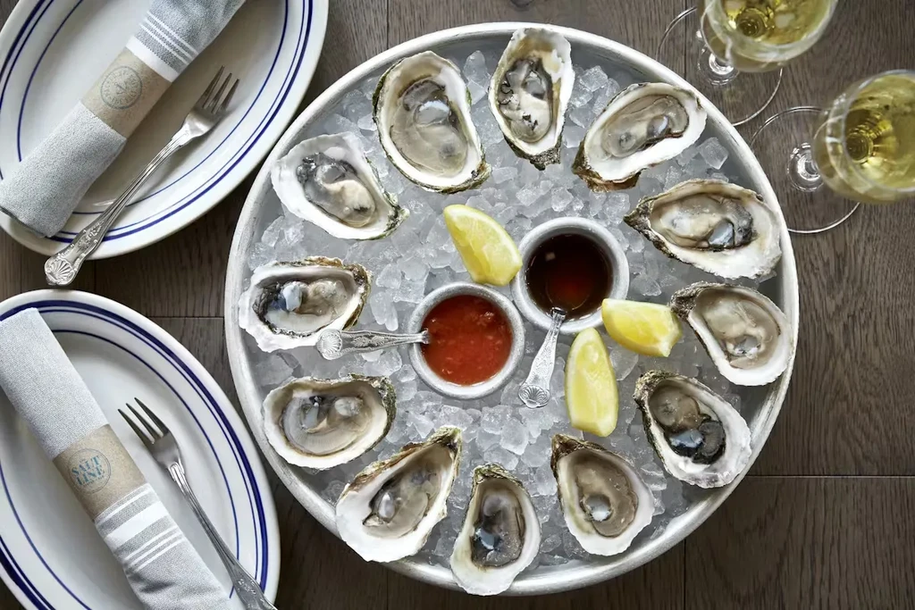 Fresh shucked oysters on ice with sauces, lemons, and wine glasses.