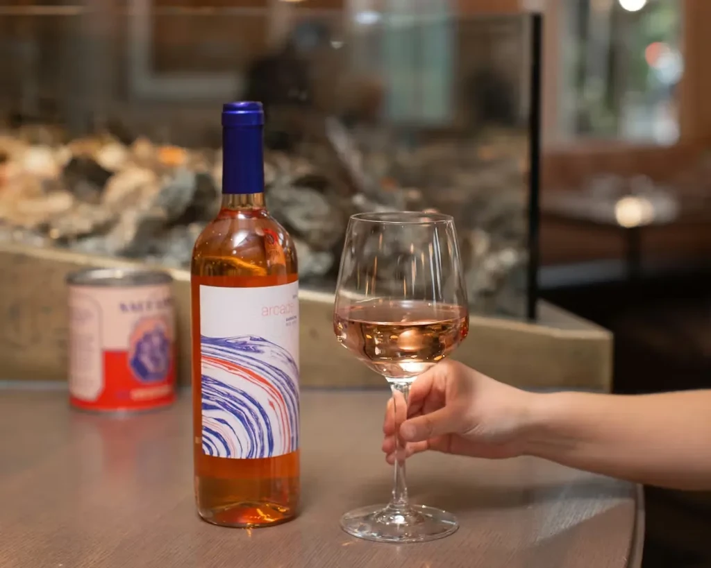Hand holds Arcade rosé wine glass and bottle at an oyster bar.