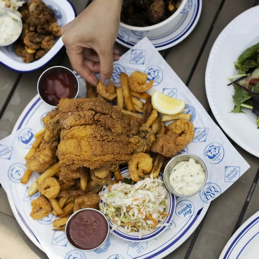 Hand picks fried shrimp from platter with fish, fries, and sauces.