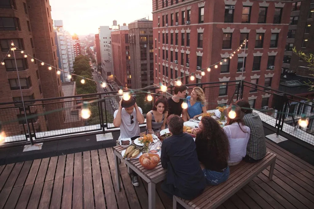 Seven people dine on a city rooftop at sunset with string lights.