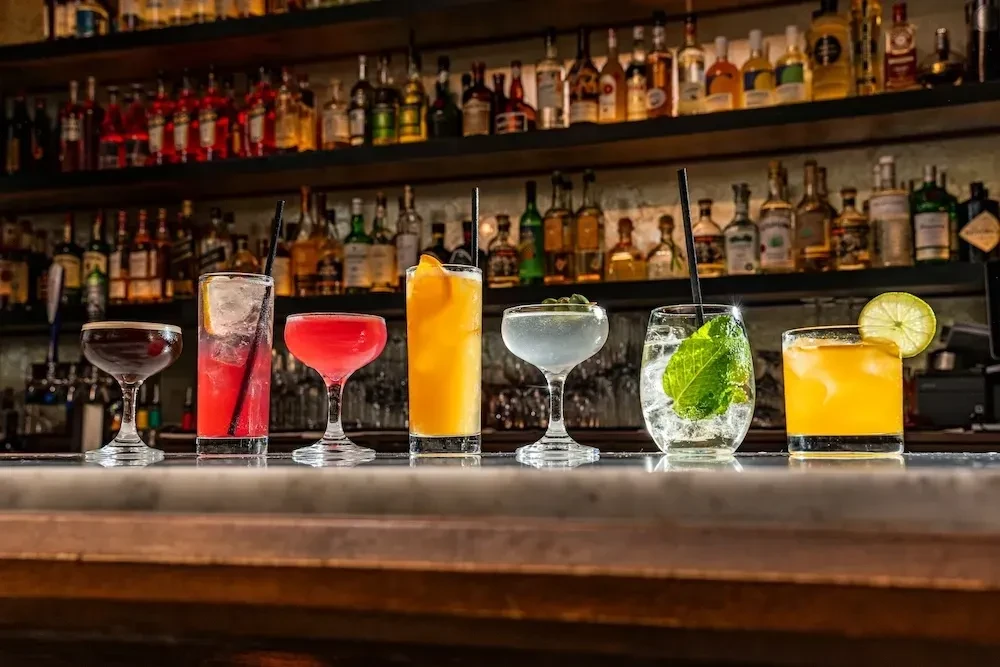Six colorful cocktails lined up on a bar with spirit bottles.