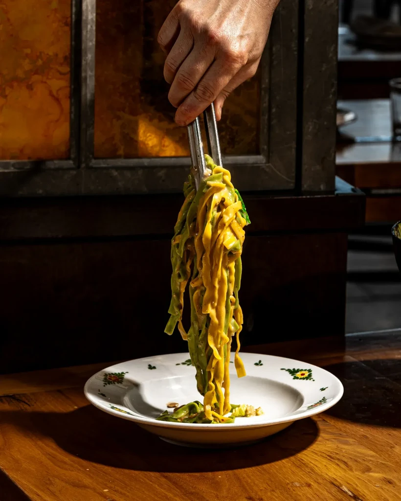 Hand uses tongs to serve green and yellow pasta with creamy sauce.