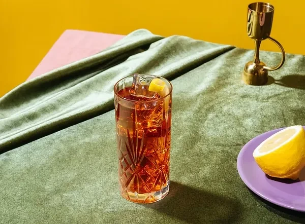 Iced reddish-brown drink, lemon peel, brass bar tool on dark velvet.