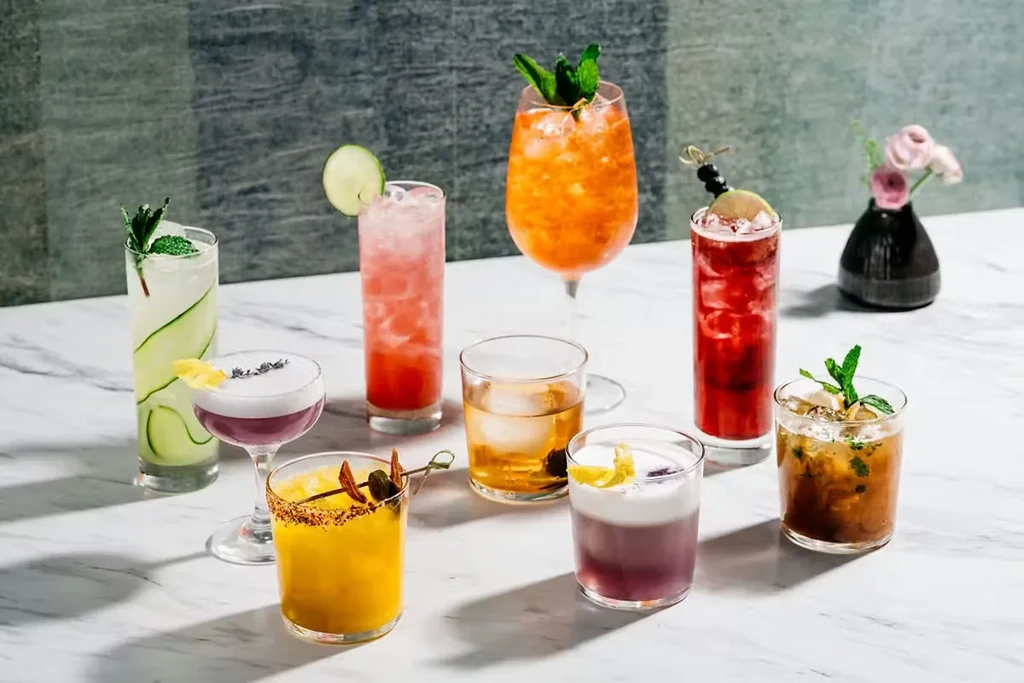 Eight colorful, garnished cocktails in various glasses on marble.