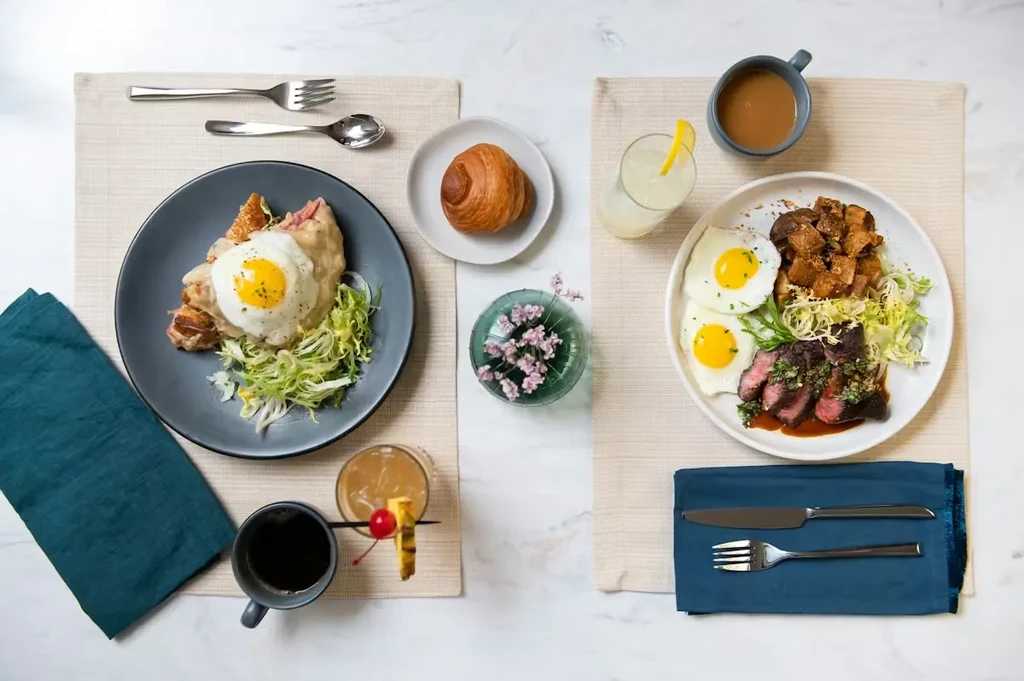 Two overhead breakfast settings with eggs, steak, croissant, and drinks on a marble table.