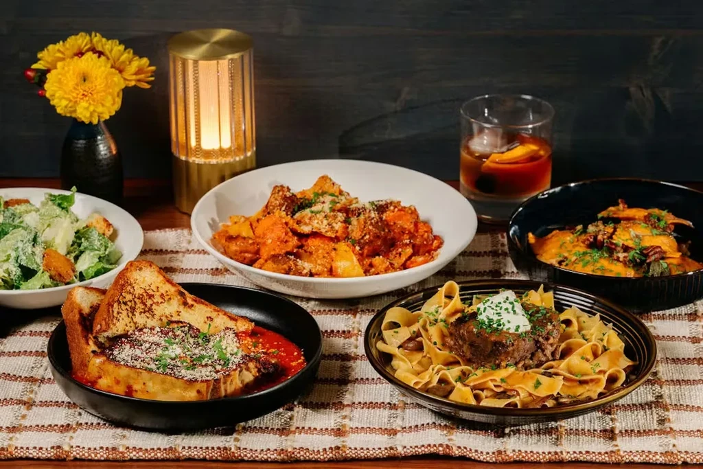 Pasta, salad, and bread dishes with a cocktail on a wooden table.