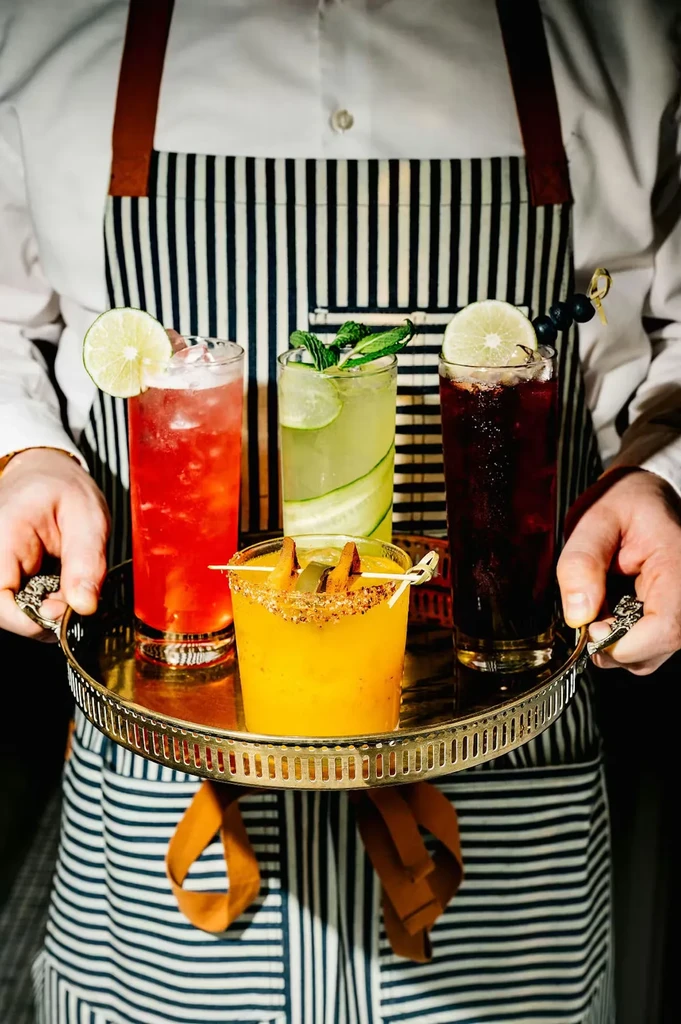 Server holding a gold tray of four colorful garnished drinks.