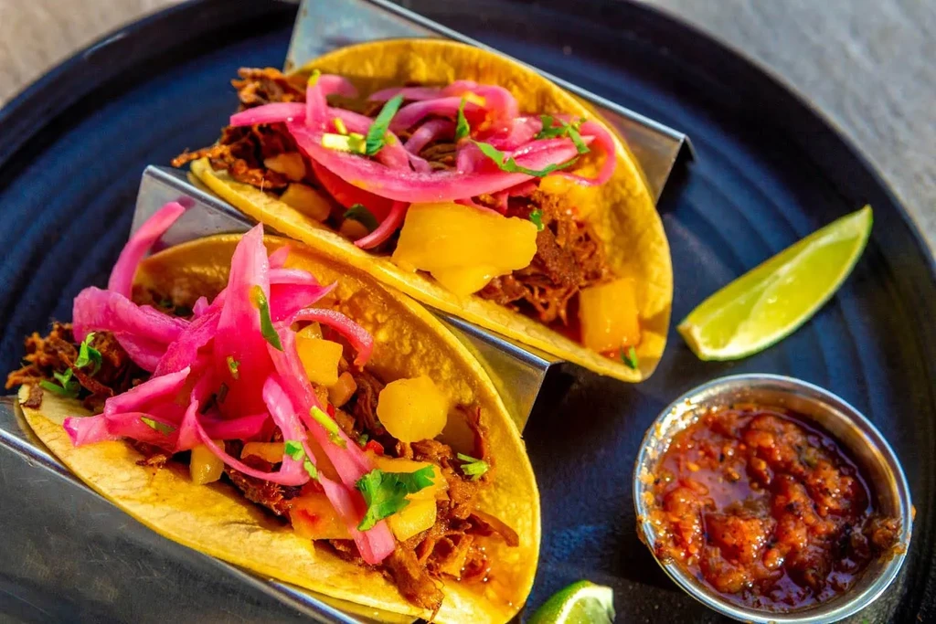 Two colorful tacos with shredded meat, pickled onions, pineapple, and cilantro.