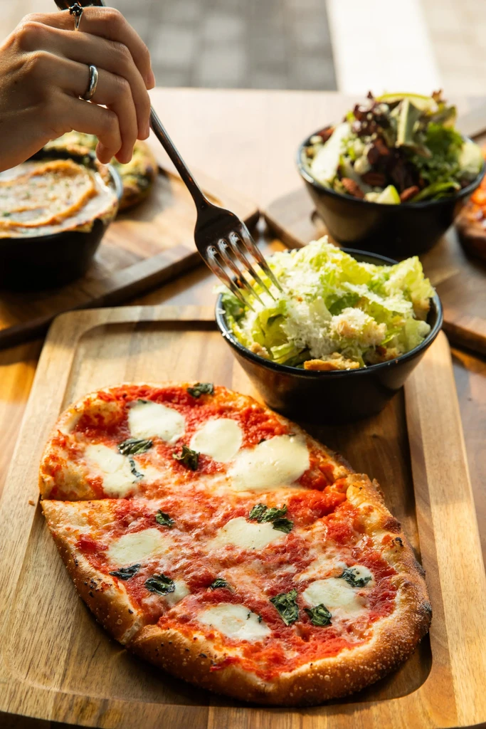 A hand forks Caesar salad next to a Margherita pizza.