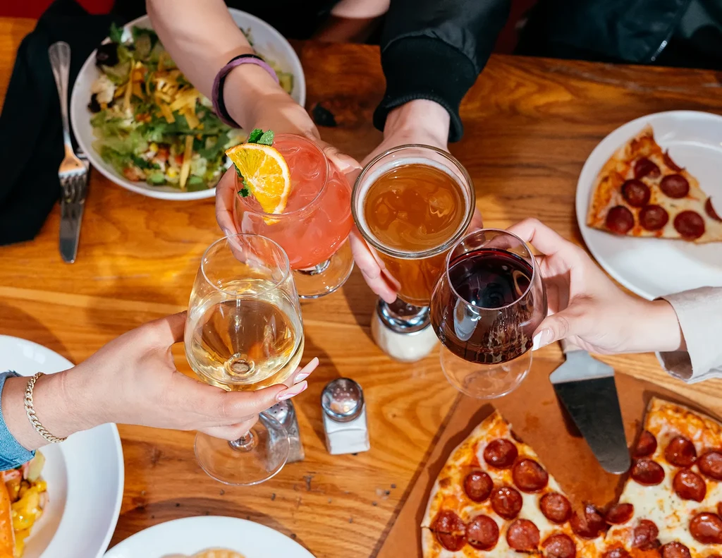 Four hands clinking glasses with various drinks, pizza, and salad.