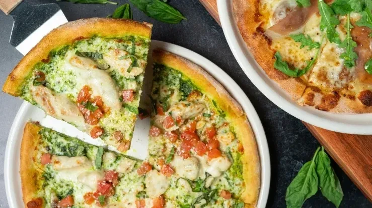 Overhead: Pesto chicken pizza with slice lifted, prosciutto pizza nearby.