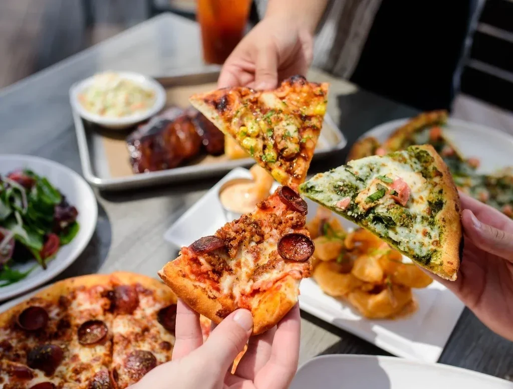 Multiple hands hold pizza slices, ribs, salad, and fried food.