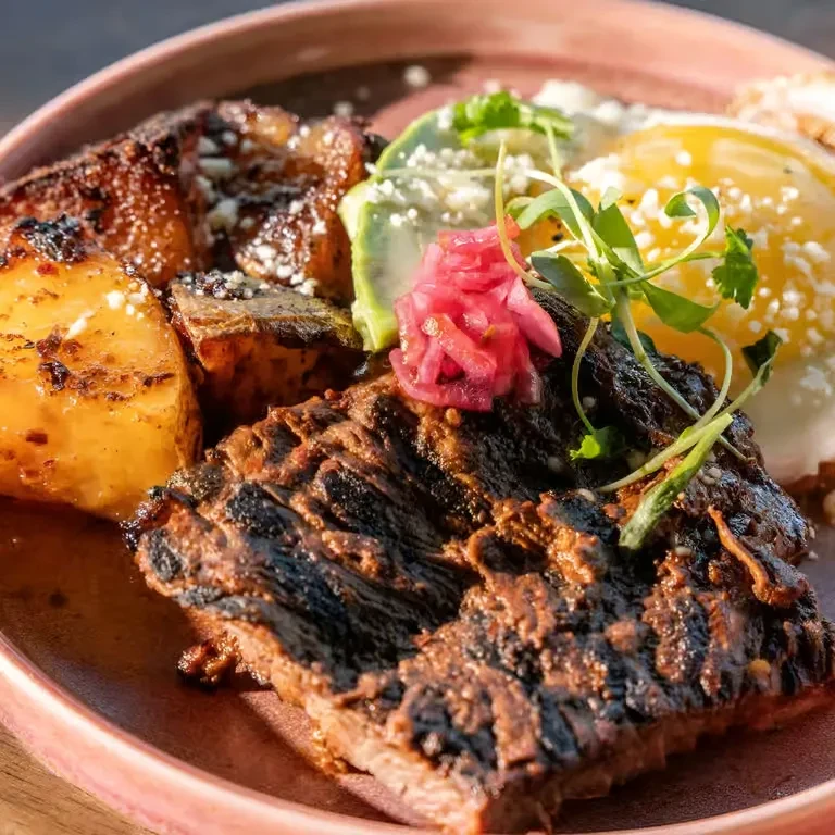 Grilled steak, fried egg, potatoes, avocado, and pickled onions on a plate.