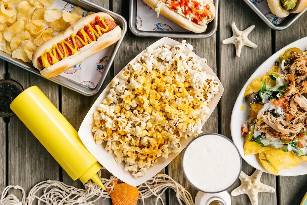 Overhead: hot dogs, loaded nachos, popcorn, beer on a decorated table.