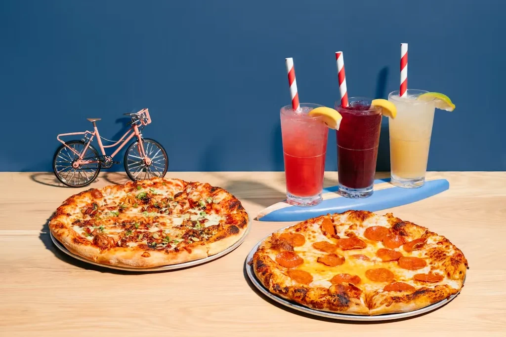 Two pizzas and three colorful, garnished drinks on a wooden surface.