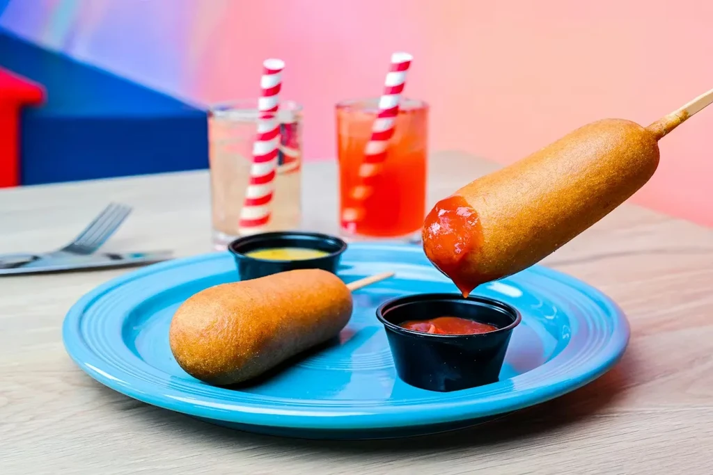 Two corn dogs, two drinks, red and yellow sauces on table.