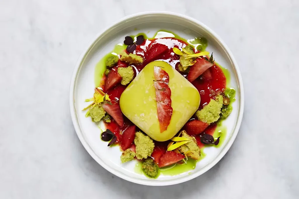 Plated light green dessert, strawberries, red sauce, green crumble, garnishes.