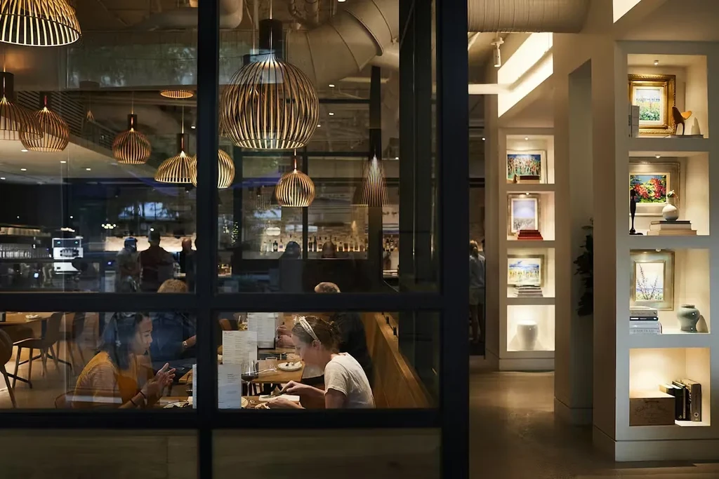 Warm-lit modern restaurant interior with diners and art-filled hallway.