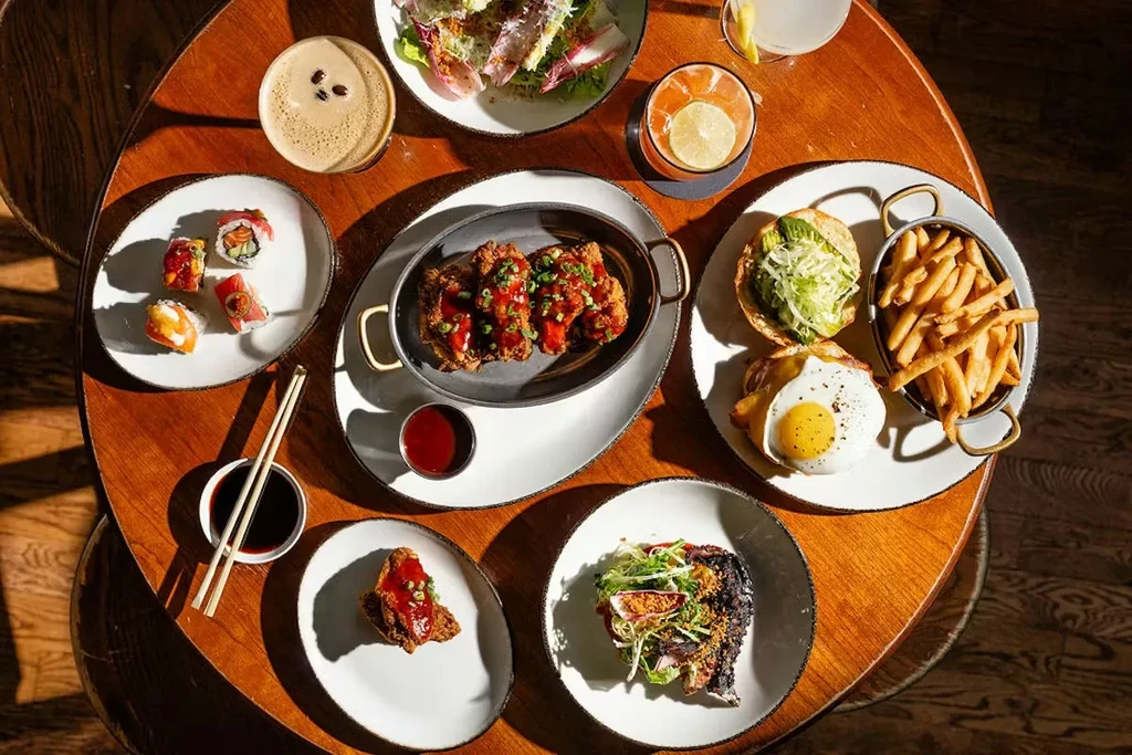 Overhead flat-lay of sushi, fried chicken, avocado bun, salad, and drinks.