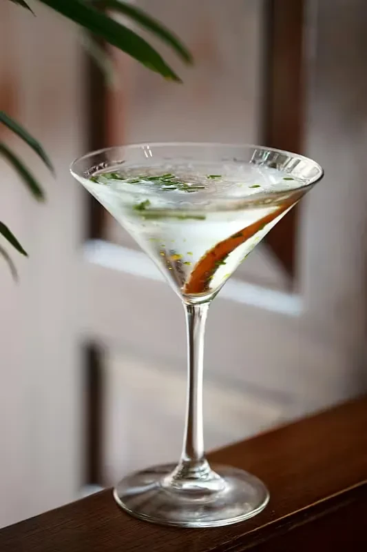 Martini glass with clear liquid, green herbs, citrus peel on wood.