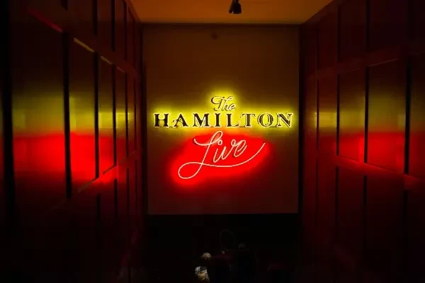 Golden "The HAMILTON" and red "Live" neon signs in a dark venue.