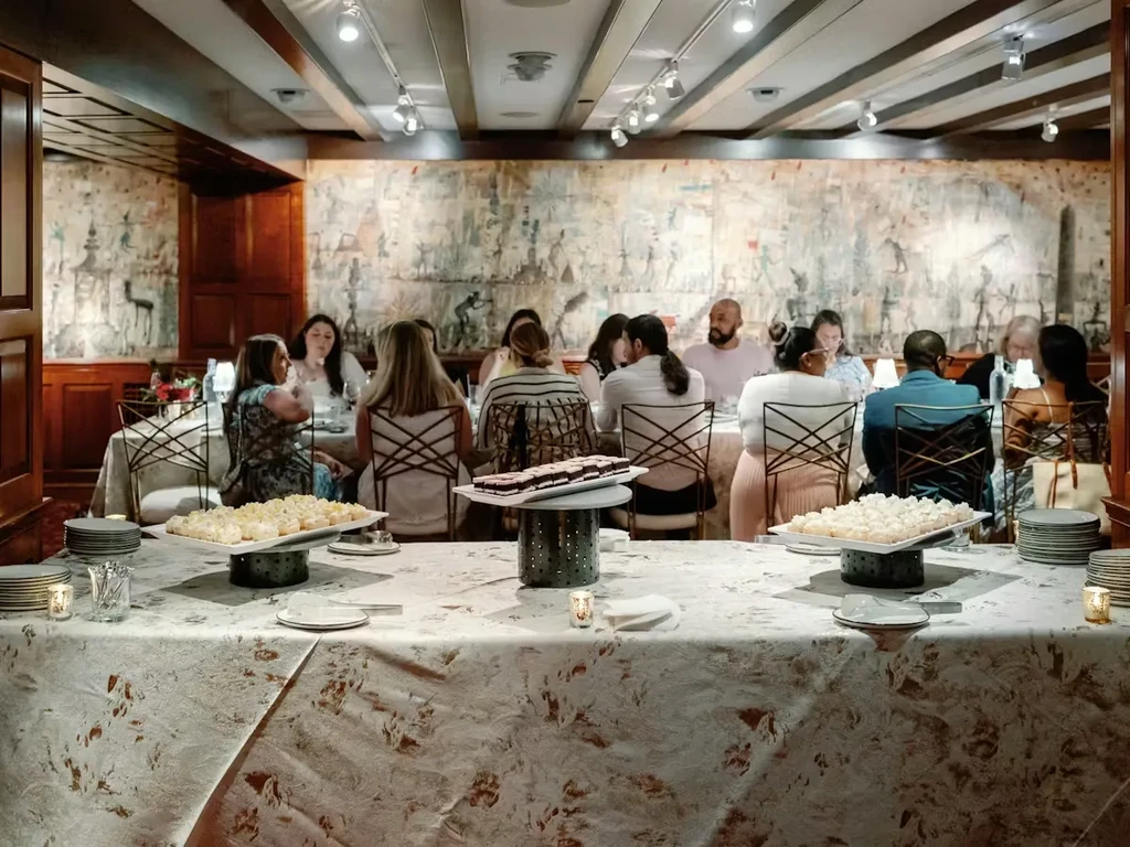 Dining room with dessert buffet, seated guests, and wall tapestry.