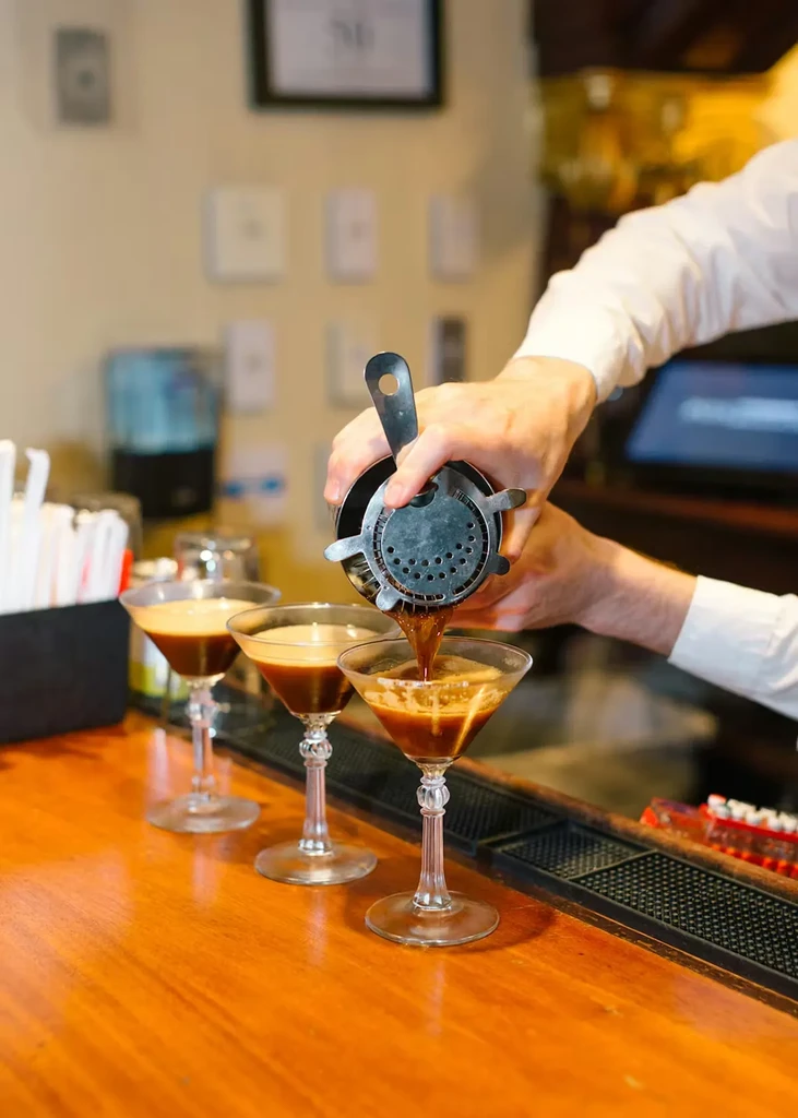 Bartender's hands pour dark liquid from a shaker into a martini glass.