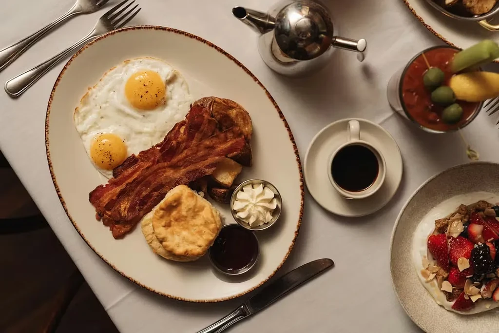 Breakfast spread of eggs, bacon, potatoes, biscuit, coffee, Bloody Mary, yogurt.