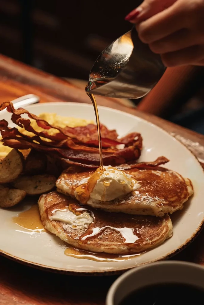 Fluffy pancakes with pouring syrup, bacon, scrambled eggs, and potato wedges.