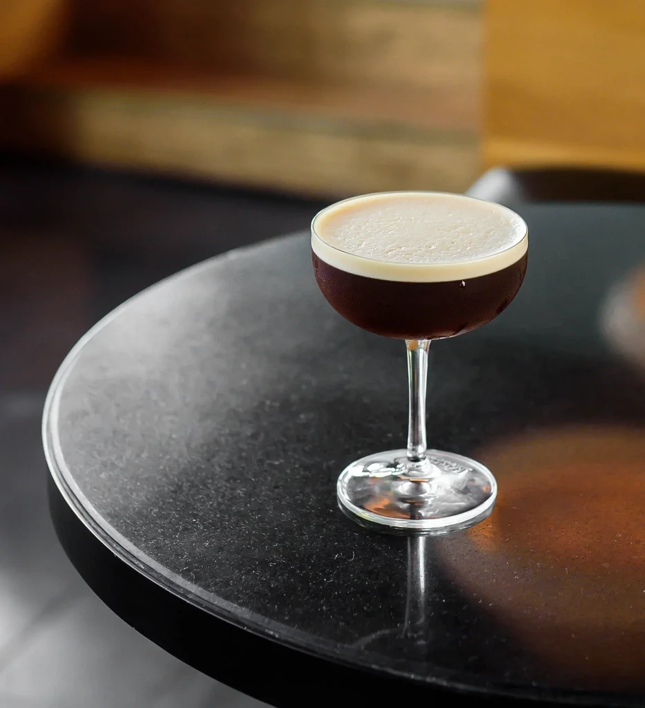 A dark brown cocktail topped with light foam on a dark table.