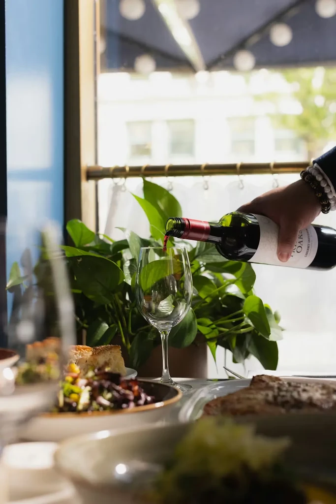 Hand pouring red wine into a glass at a restaurant table with food.