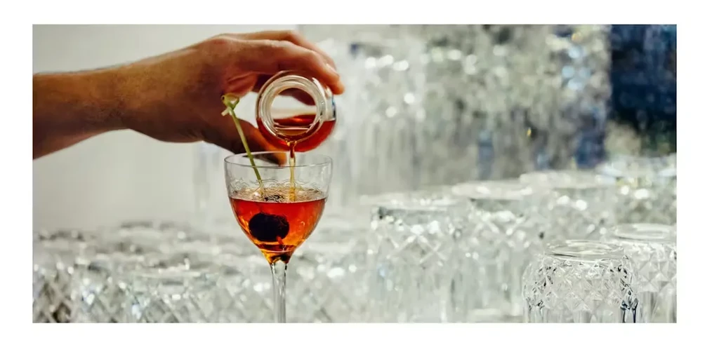 Bartender's hand pours amber liquid into a cocktail glass with a cherry.