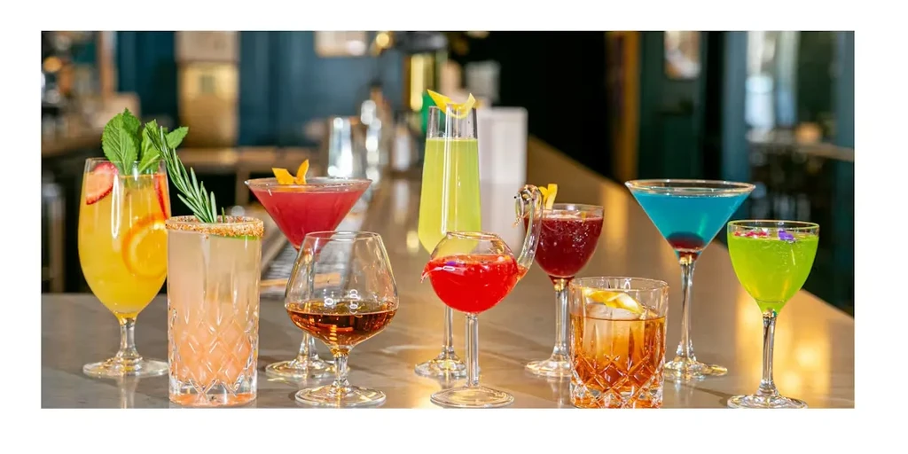 Nine colorful cocktails with garnishes in distinct glasses on a bar.
