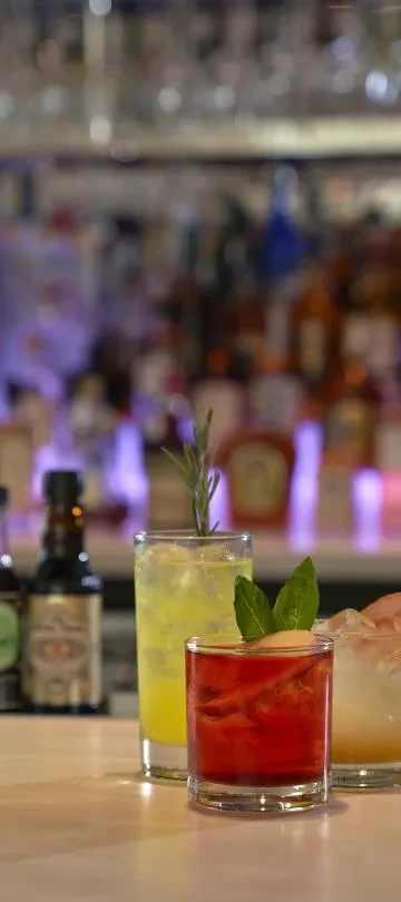 Two vibrant, garnished cocktails, yellow and red, on a polished bar.