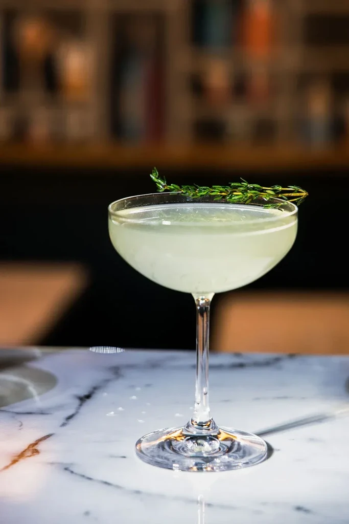 Greenish-yellow cocktail in coupe glass with herb garnish on marble.