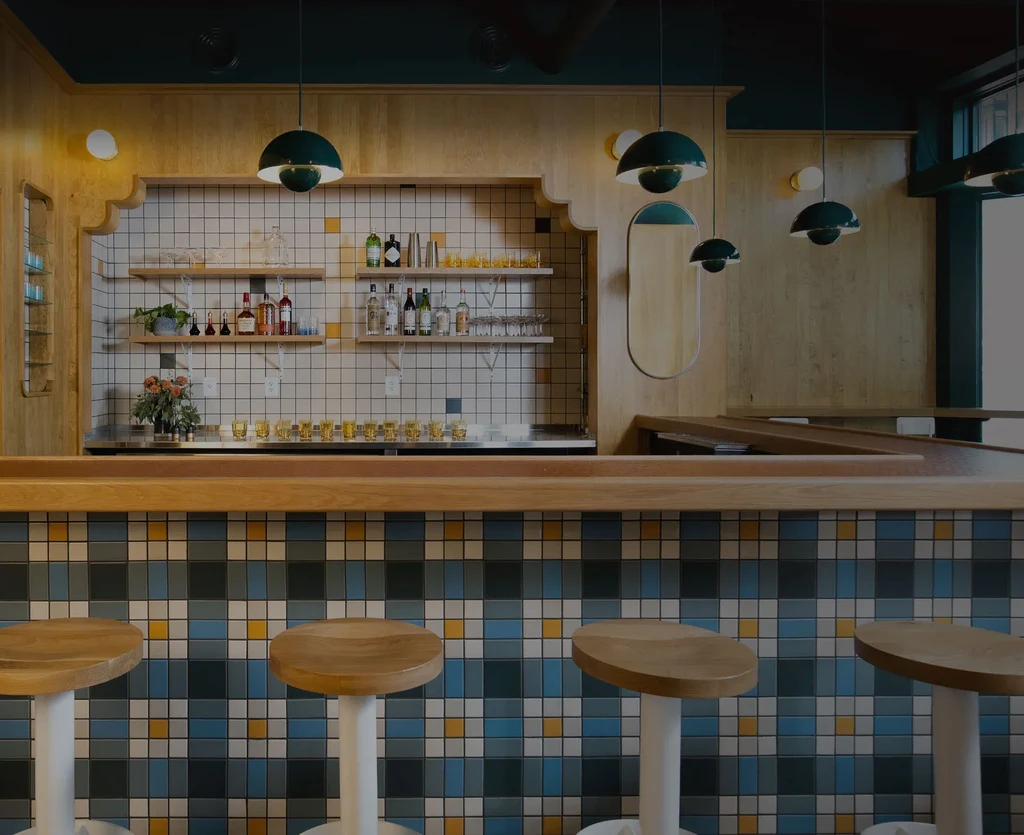Modern bar with colorful checkered tiles, wooden stools, and arched shelves.