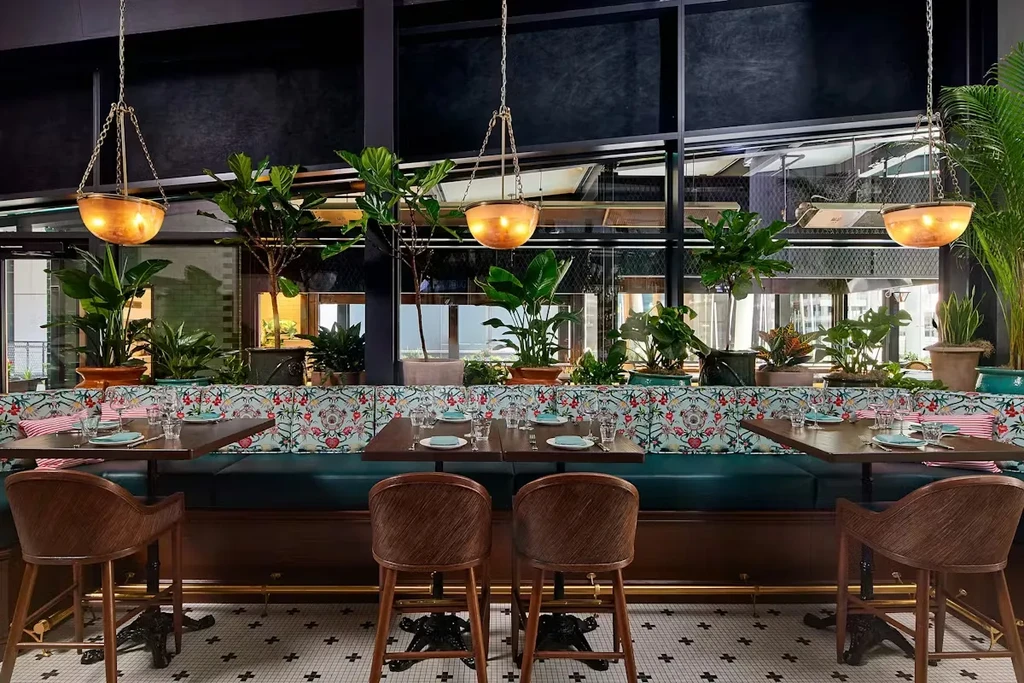 Empty restaurant interior: floral booth, wooden tables, green plants, pendant lights.