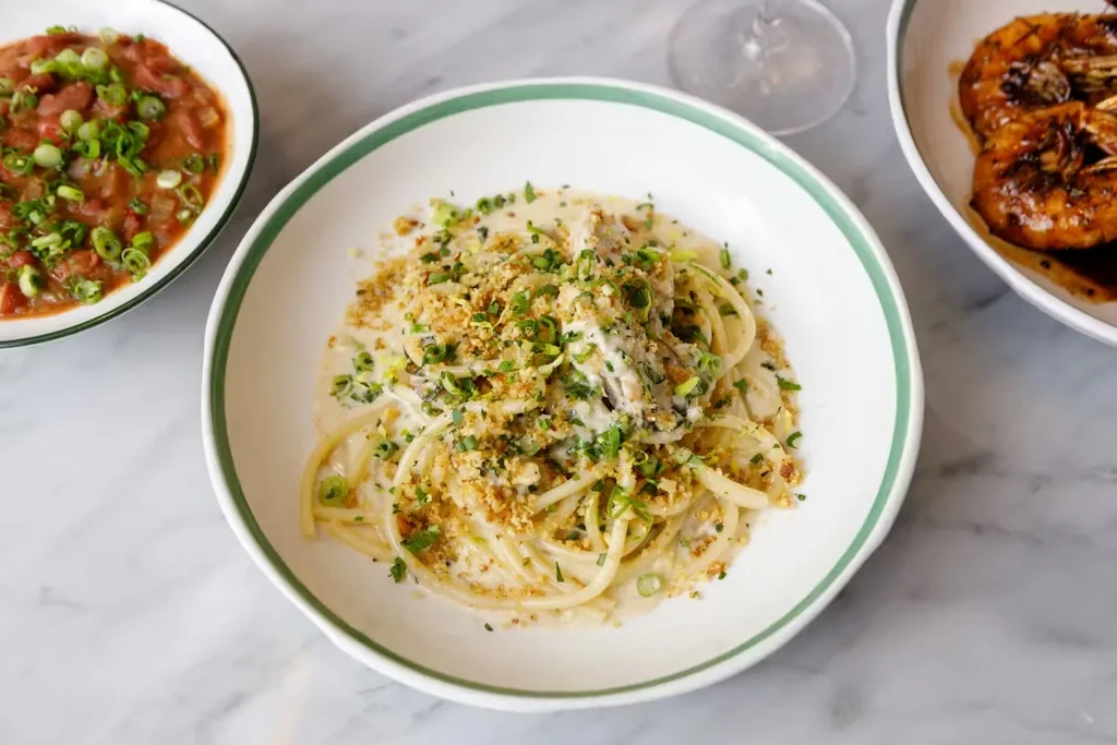 Top-down: Creamy spaghetti with herbs, breadcrumbs, and sides on marble table.