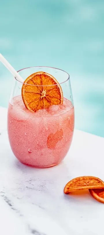 Vibrant pink frozen drink in glass, garnished with dried orange and straw.
